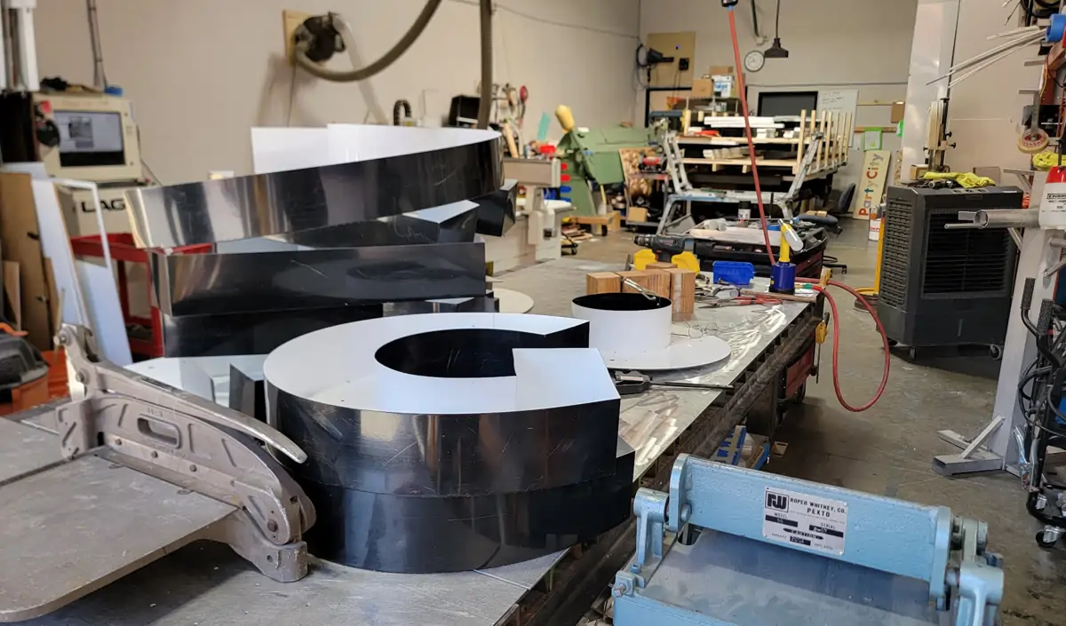 Workshop interior showing large channel‑letter components (including a glossy black and white "C"), fabrication tools, table saws, clamps, and partially assembled signage elements.