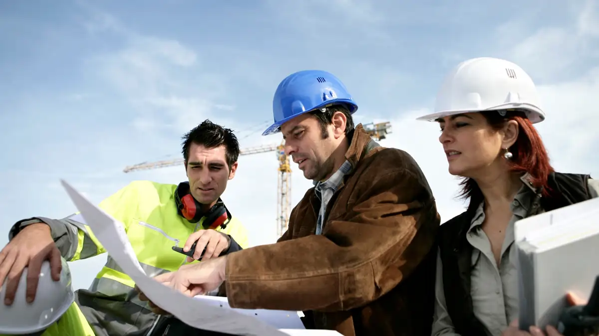Three project staff at a construction site reviewing plans; two wearing hard hats and one in a hi‑vis jacket with ear protection; cranes and active work in background.