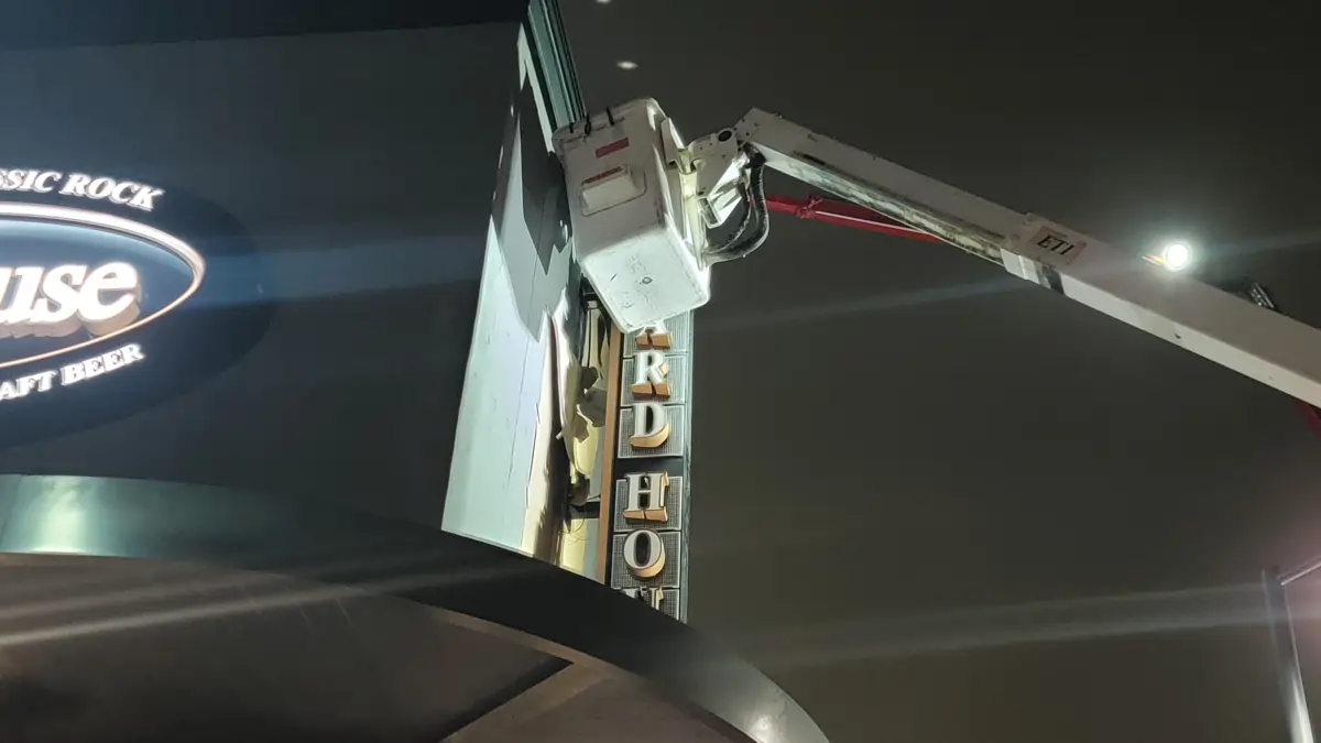 Certified crew technician in a bucket lift installing an illuminated vertical YardHouse sign at night with ETI lift and lit oval “Classic Rock Craft Beer” sign on the building.
